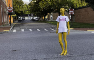 gold painted mannequinn stands in middle of road wearing white shirt with pink womens lives matter flag