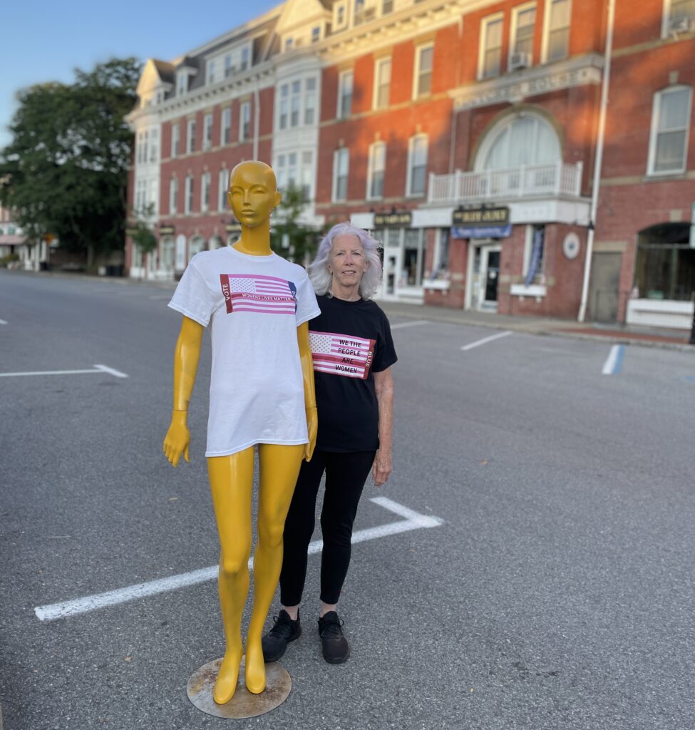 woman dressed in black wearing pink lives matter flag shirt standing in road with gold painted manniquin with similar shirt