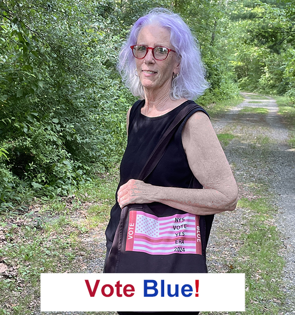 woman with purplish hair standing on wooded path carries black tote bag with pink flag and message N Y S vote yes E R A